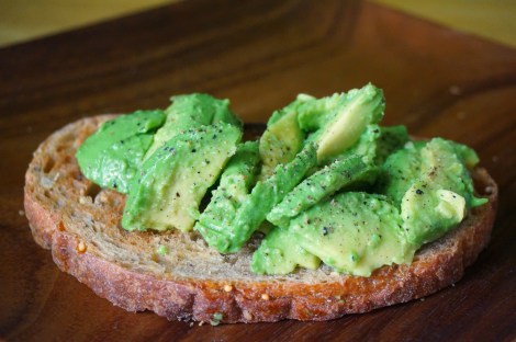 Avocado toast sits on wooden plate, pale green scallops of avocado piled high on a lightly toasted slice of a multigrain boule.