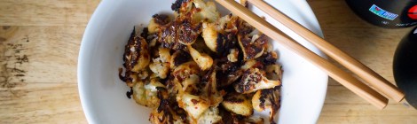 A small white bowl of spicy roasted cauliflower, with a pair of chopsticks balanced on its edge. In the background we see the edges of bottles of chile paste, soy sauce, and sesame oil.