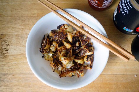 A small white bowl of spicy roasted cauliflower, with a pair of chopsticks balanced on its edge. In the background we see the edges of bottles of chile paste, soy sauce, and sesame oil.
