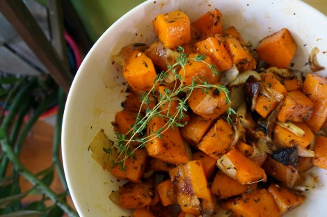 A overhead shot of chunks of honey-vinegar butternut squash rest, piled in a white bowls, with a decorative sprig of fresh thyme on top. In the background we see dark green houseplants.