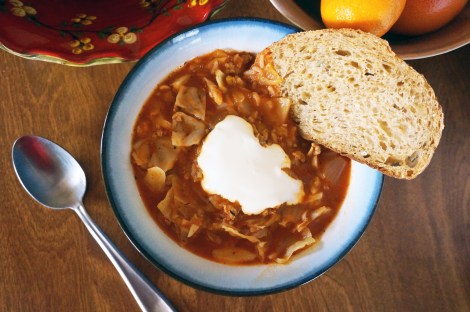 Stuffed cabbage soup, in an overhead shot. A slice of multigrain bread rests on the rim of the bowl, and a big dollop of sour cream floats in the middle of the brick-red soup. Bowls of produce peek in at the edges of the image.