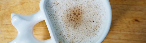 Cardamom coffee in a mug whose handle is modeled after a human vertebra. A layer of bubbly froth covers the top of the mug, with a warm brown smudge in the center where the coffee was poured.