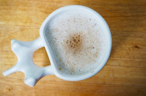 Cardamom coffee in a mug whose handle is modeled after a human vertebra. A layer of bubbly froth covers the top of the mug, with a warm brown smudge in the center where the coffee was poured.