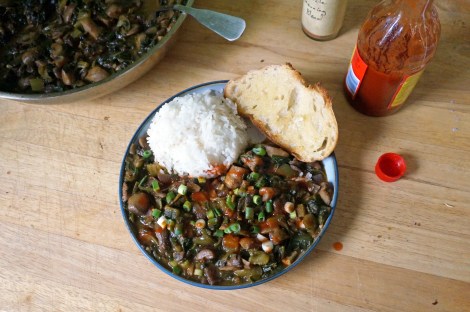 A plate of smothered mushrooms with spinach, accompanied by white rice and buttered bread, garnished with chopped green onions and hot sauce. A skillet of the dish rests at the edge of the picture, and bottles of Creole seasoning and hot sauce sit near the plate