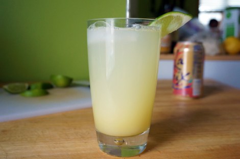 A glass of ginger beer, garnished with a wedge of lime at a jaunty angle, sits on a wooden countertop. In the background, wedges of lime on a cutting board, and a pink and orange can of La Croix sparkling water.