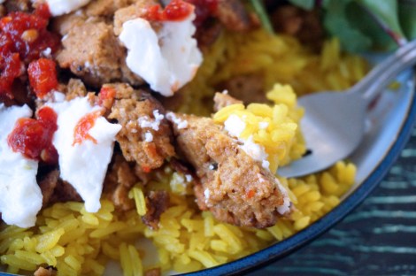 In a close-up, a chunk of street-cart seitan sits with a bite of bright-yellow on a fork, the whole bite dappled with white garlic sauce and vibrant red chile paste. In the background, more seitan and rice, and the edge of some leaves of salad.