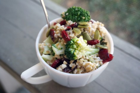A white teacup rests on a grey wooden rail, filled to near-overflowing with garlicky oat and broccoli salad.