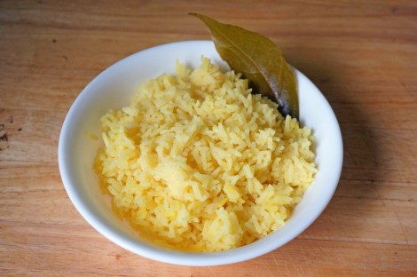 Pale yellow bay leaf rice, served in a small white bowl sitting on a wooden countertop. An olive-colored cooked bay leaf is tucked in along the edge of the bowl.