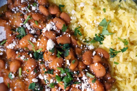Extreme closeup on pinto beans stewed with cilantro and bay: orangey-pink beans and light yellow rice fill the entire frame, all festooned with chopped herbs and crumbled cheese