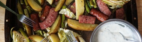A dark brown rimmed baking sheet filled with pan-roasted asparagus, cabbage, potato, and smoked sausage, with a big bowl of herb-flecked yogurt sauce in the corner, and a whole bunch of forks laid around, ready to dig in