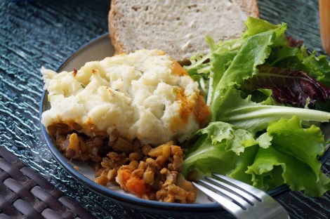 A hefty scoop of bacon and lentil shepherd's takes up most of a plate on a glass patio table, the reddish-brown lentil filling spilling out from beneath ivory potatoes. Buttered bread and a green salad nestle close to the main dish, with the tines of a fork protruding in from the lower edge of the fram.