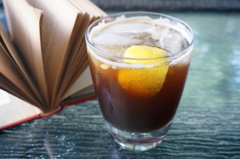 A simple rocks glass sits on a glass tabletop, filled with a fizzing, dark brown, iced drink with a wedge of lemon floating at the near side of the glass. Behind the glass, a book lies open on the table, pages spreading like a fan.