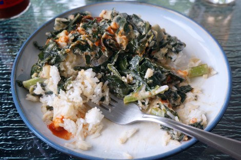 A plate of collards with coconut and peanut butter, drizzled with hot sauce, sits partially eaten, a fork nestled among the rice grains and dark green leaves.