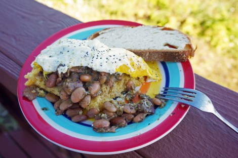 A thick slice of cornbread, covered in beans and topped with a fried egg, sits on a brightly-colored plate on a porch rail.