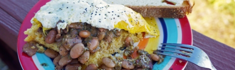 A thick slice of cornbread, covered in beans and topped with a fried egg, sits on a brightly-colored plate on a porch rail.