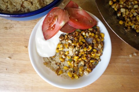 A big scoop of quinoa in a white bowl, topped with bright yellow corn spotted with brown and dressed with a dollop of yogurt and a few slices of bright red tomato. At the edges of the picture, a serving bowl of quinoa and a wok full of more charred corn.