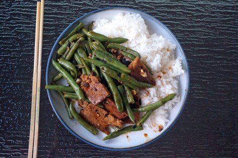 On a glass table, a blue-rimmed plate, with a pair of chopsticks laid to the left. On the plate, a mound of fluffy white rice, half-covered with wrinkled green beans and slices of dark brown pan-crisped seitan, all dressed lightly in a brown sauce and flecked with black, beige, and red-brown bits of seasoning.