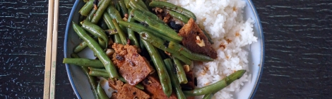 On a glass table, a blue-rimmed plate, with a pair of chopsticks laid to the left. On the plate, a mound of fluffy white rice, half-covered with wrinkled green beans and slices of dark brown pan-crisped seitan, all dressed lightly in a brown sauce and flecked with black, beige, and red-brown bits of seasoning.