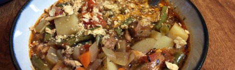 A bowl of chicken stew with warm, wintry spices, chunks of red-skinned potatoes, chicken, carrot, and green beans bobbing in an orange-brown broth. A stainless steel spoon handle catches the light, and a scattering of crushed tortilla chips garnishes the bowl. In the background, a dark blue serving bowl awaits.