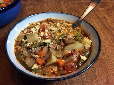 A bowl of chicken stew with warm, wintry spices, chunks of golden-skinned potatoes, chicken, carrot, and green beans bobbing in an orange-brown broth. A stainless steel spoon handle catches the light, and a scattering of crushed tortilla chips garnishes the bowl. In the background, a dark blue serving bowl awaits.