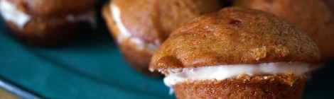 In focus in the foreground, a tawny brown curried pumpkin mini-muffin, split and spread with cream cheese frosting in the middle. Behind it, out of focus, part of a mound of those muffins on a turquoise enamel plate.