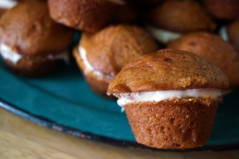 In focus in the foreground, a tawny brown curried pumpkin mini-muffin, split and spread with cream cheese frosting in the middle. Behind it, out of focus, part of a mound of those muffins on a turquoise enamel plate.