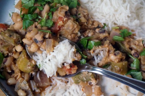 Closeup on a spoon of black-eyed pea and okra curry, with rice, sitting in the midst of the larger plateful, the delicate white grains of rice contrasting with the hearty thickness of brown, red, olive, and green curry ingredients.