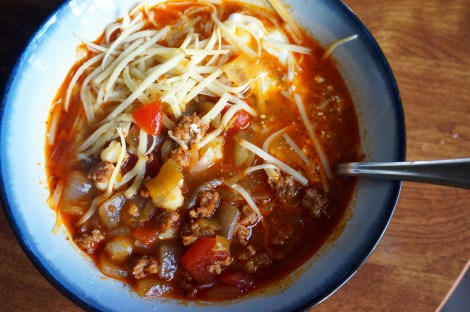 Close-up on a bowl of quick and lazy pozole, deep brick-red broth studded with brown chorizo, bright red tomatoes, yellow-green chiles, and pale yellow hominy. A small pile of cheese sits near a dollop of sour cream near the upper edge of the bowl.