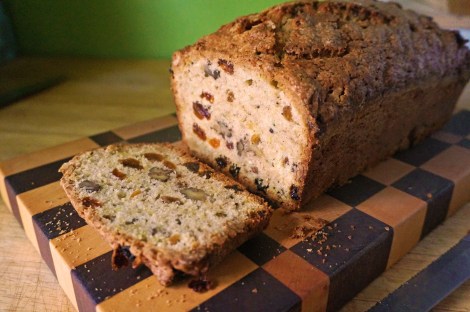 A slice of zucchini bread lies next to the rest of the loaf, all resting on a checkerboard wooden cutting block, which itself contrasts with the blonde wood counter and a bright green painted wall.